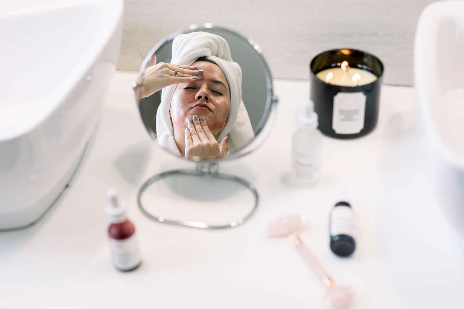 A woman with a towel wrapped around her head applies skincare to her face while looking into a small mirror, with candles, serums, and a facial roller on the counter.